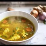 A bowl of steaming Immune Boosting Broth with chicken, carrots, and herbs. Ginger and garlic are in the background.