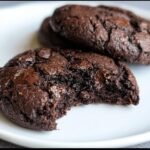 Two Keto Flourless Fudge Cookies on a plate, one with a bite taken out, showing the fudgy interior.