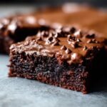 A slice of Secretly Healthy Zucchini Brownies with chocolate frosting and mini chocolate chips on a gray surface.