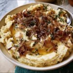 A bowl of creamy mashed potatoes topped with caramelized onions and fresh thyme.