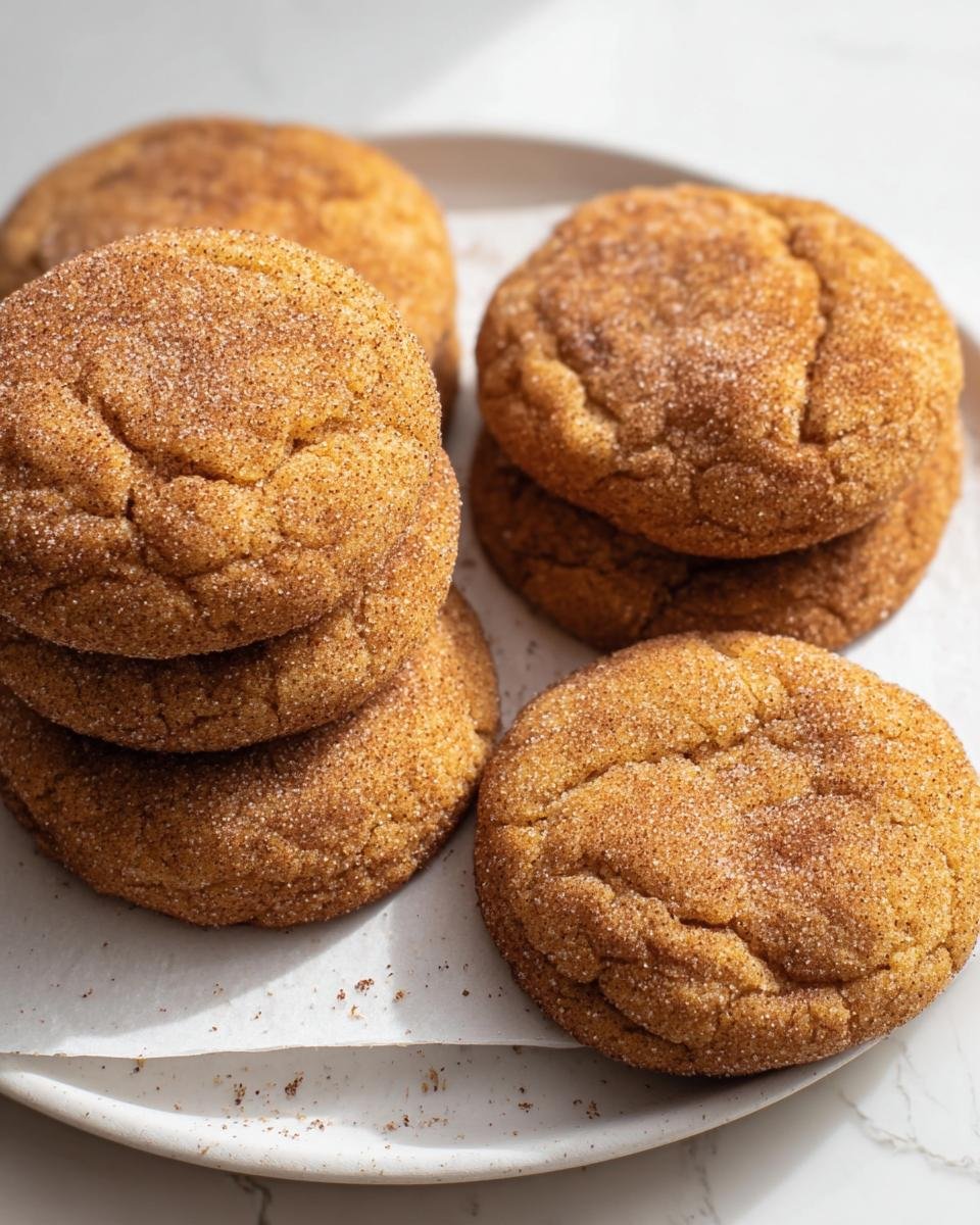 Several chewy pumpkin snickerdoodles coated in cinnamon sugar, stacked on a white plate.