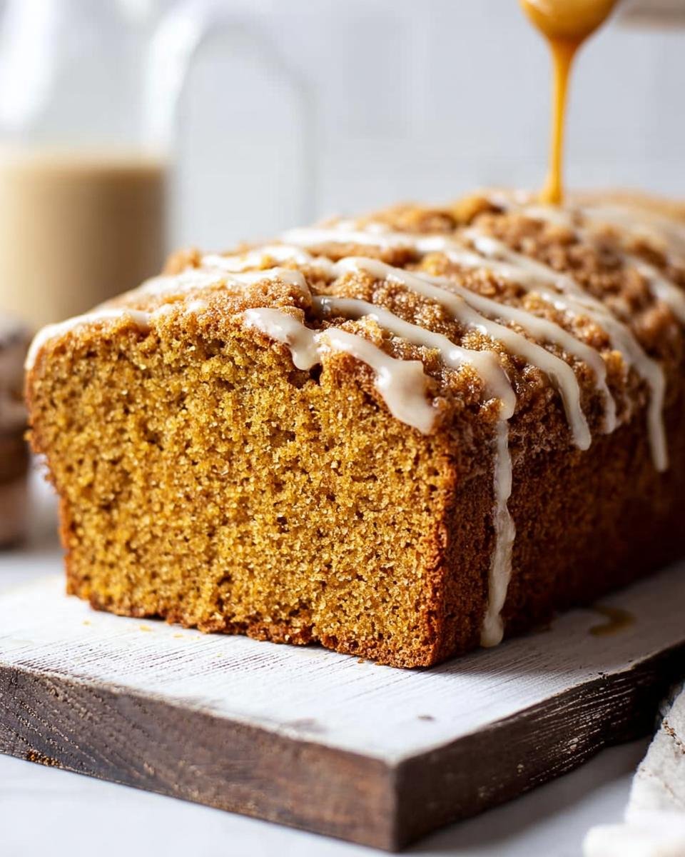 A loaf of Pumpkin Bread with Maple Glaze being drizzled on top, sitting on a wooden board.