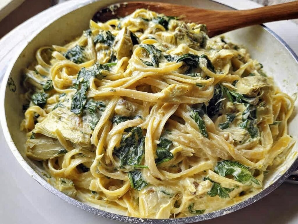 Close-up of fettuccine coated in rich sauce with spinach and artichoke hearts in a skillet, featuring Creamy Spinach Artichoke Pasta Comfort Food.