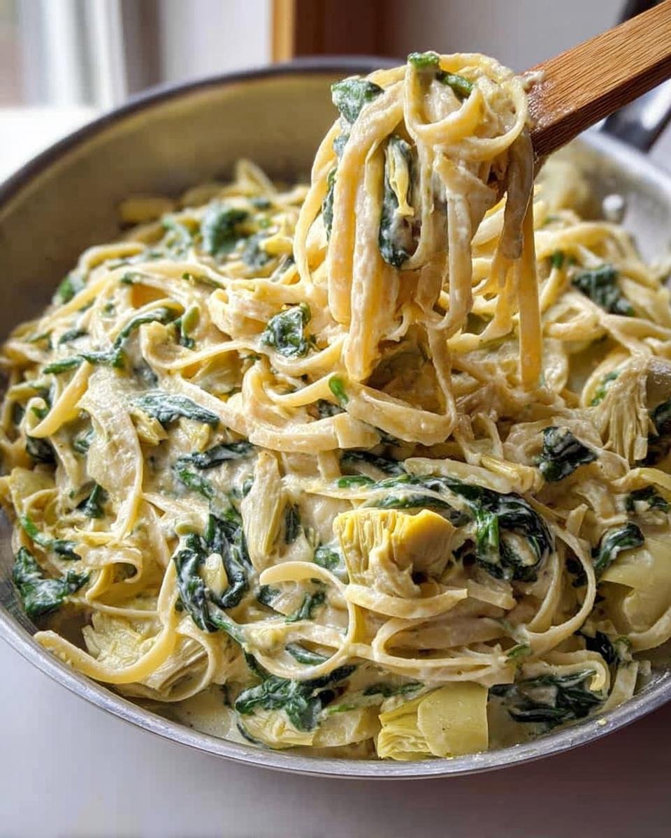 A wooden spoon lifts fettuccine coated in a rich sauce with spinach and artichoke hearts from a pan of Creamy Spinach Artichoke Pasta.