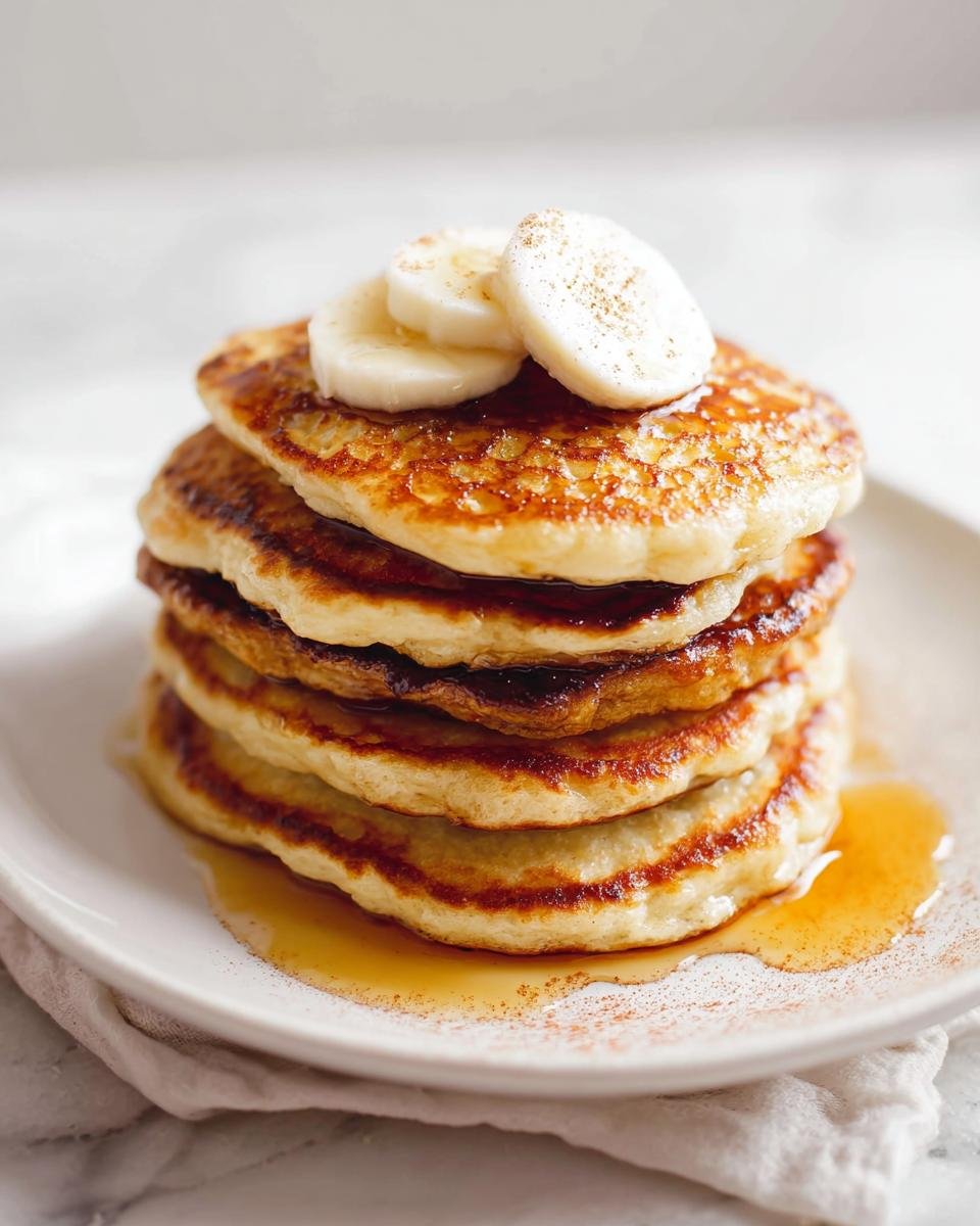 A tall stack of fluffy Protein Pancakes Fluffy drizzled with syrup and topped with banana slices.