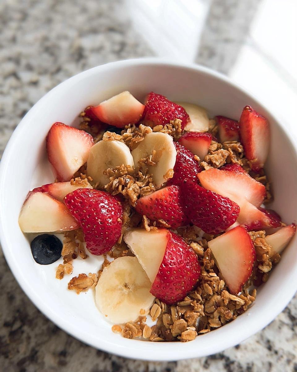 A close-up of a Greek Yogurt Fruit Breakfast Bowl topped with fresh strawberries, banana slices, and granola.