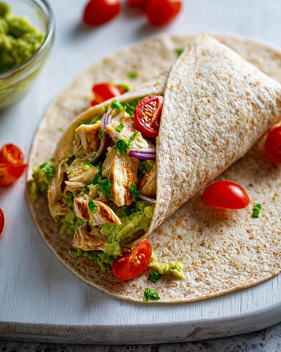 A healthy chicken avocado wrap filled with shredded chicken, mashed avocado, red onion, and cherry tomatoes.