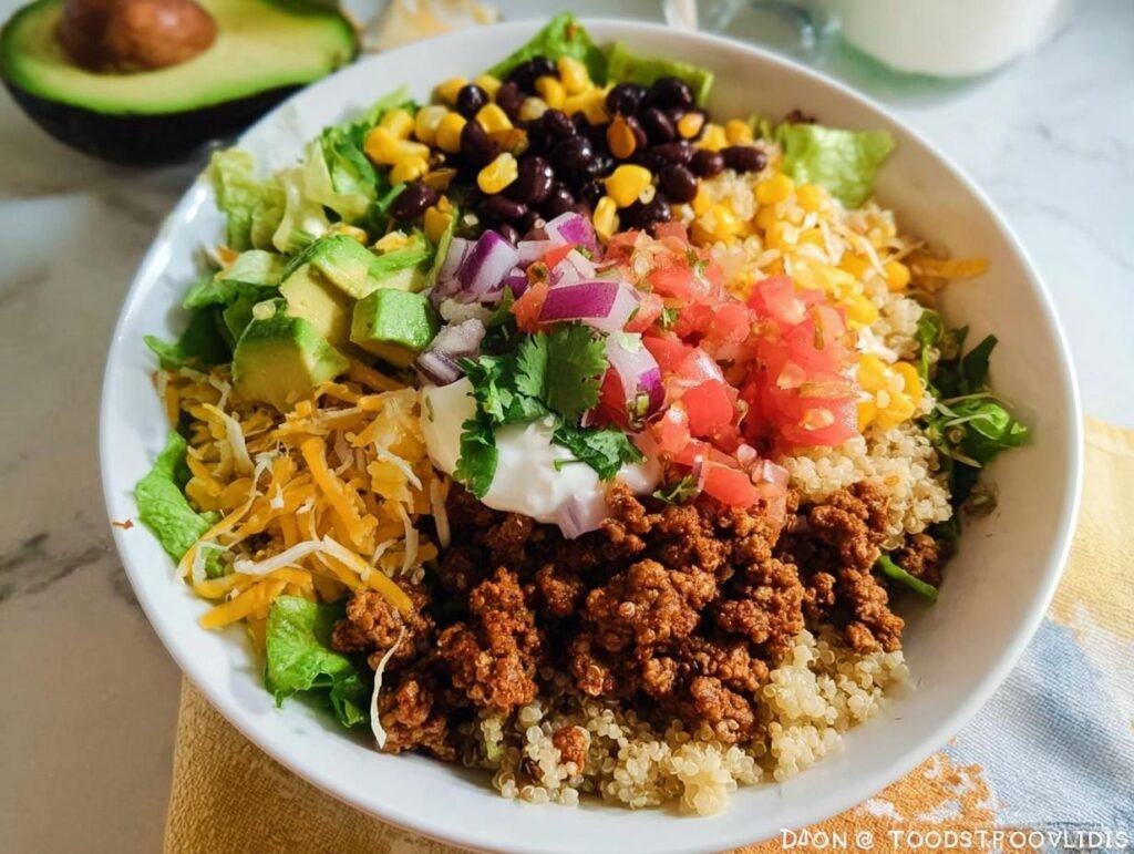 A vibrant and healthy taco bowl filled with seasoned ground meat, quinoa, black beans, corn, lettuce, avocado, pico de gallo, and sour cream.