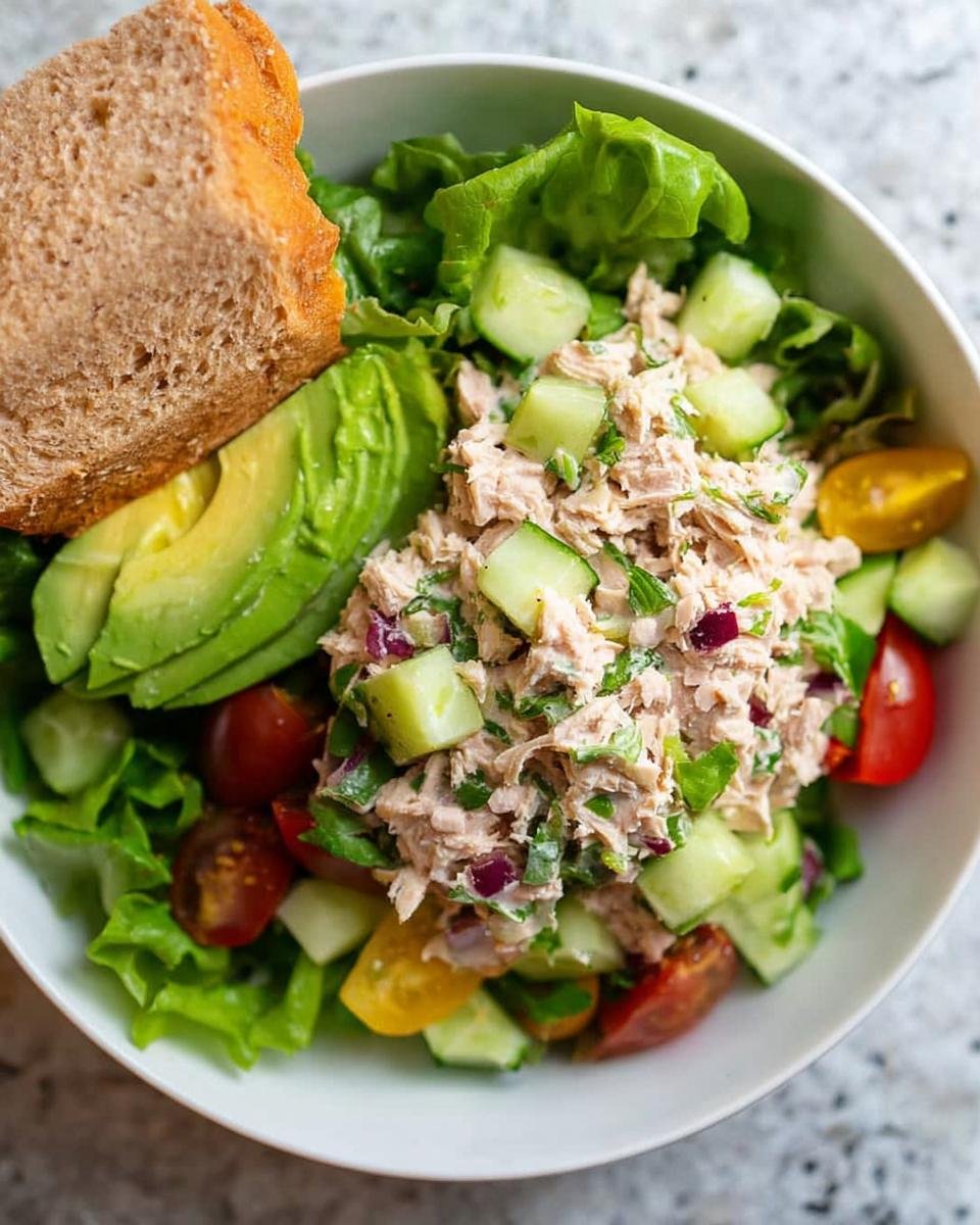 A close-up of a Healthy Tuna Salad Bowl with tuna, avocado, cucumber, tomatoes, lettuce, and a slice of bread.