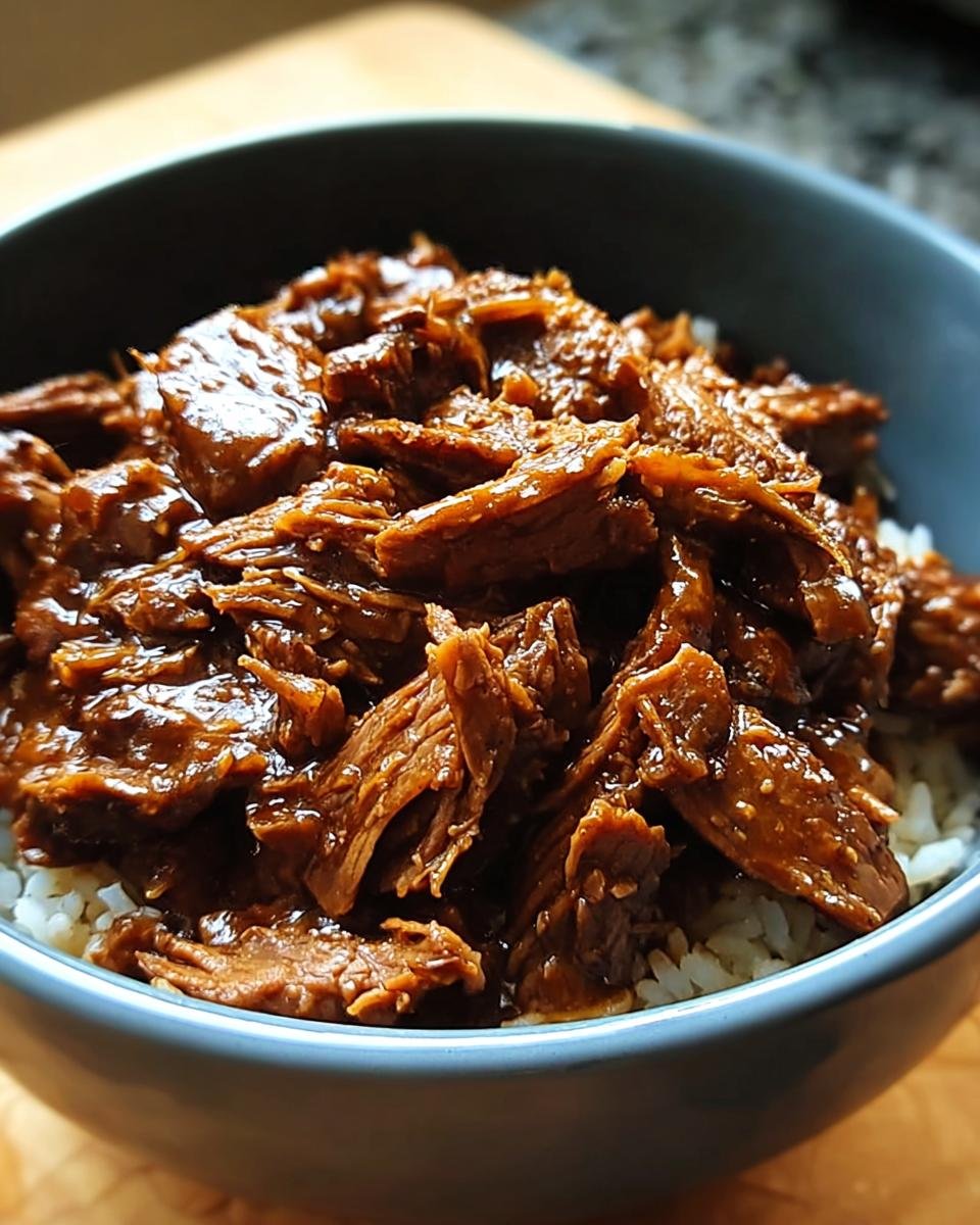 Close-up of tender Slow Cooker Korean Beef piled high over white rice in a blue bowl.