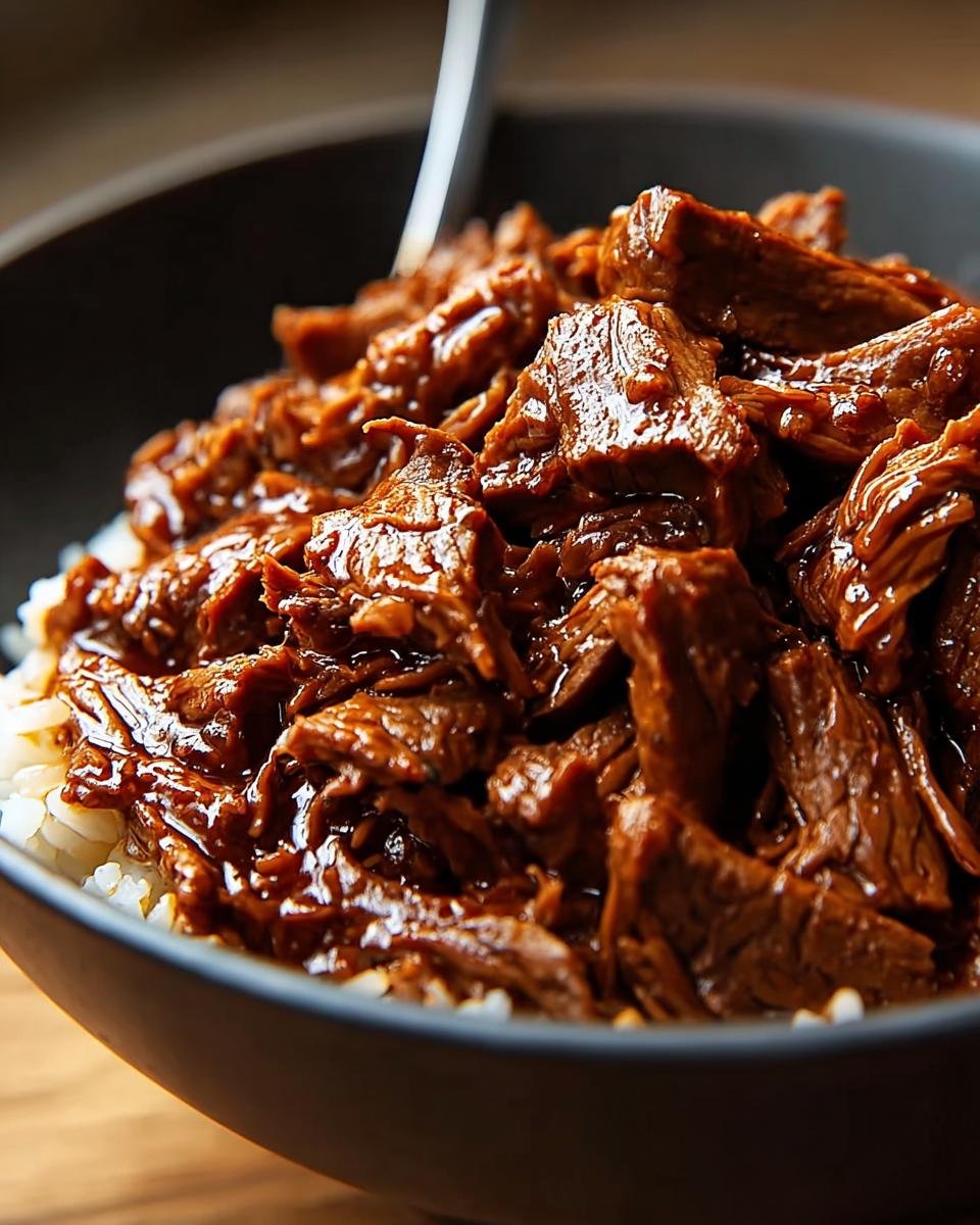 Close-up of tender Slow Cooker Korean Beef piled high over fluffy white rice in a dark bowl.