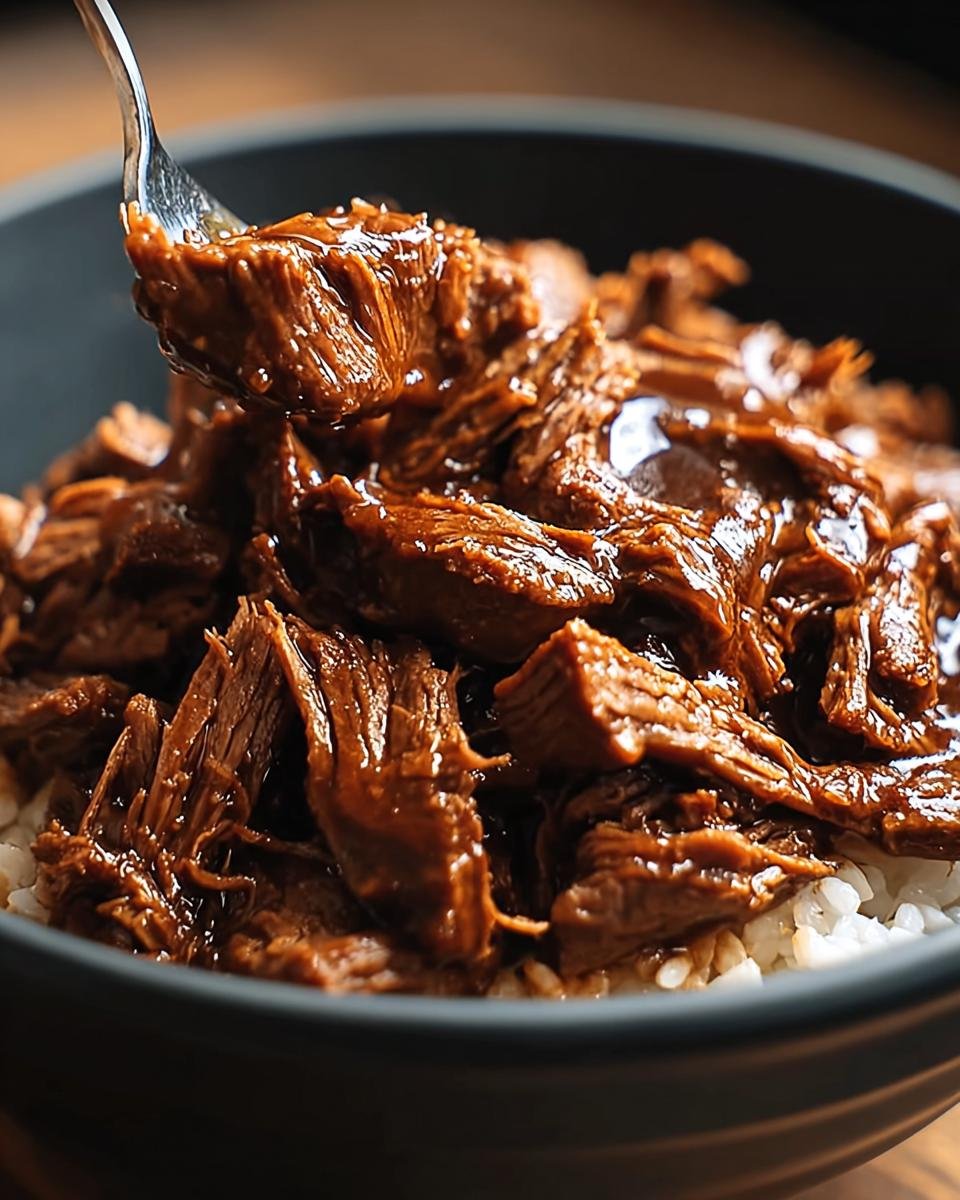A fork lifting tender, shredded Slow Cooker Korean Beef coated in a glossy sauce, served over white rice in a dark bowl.